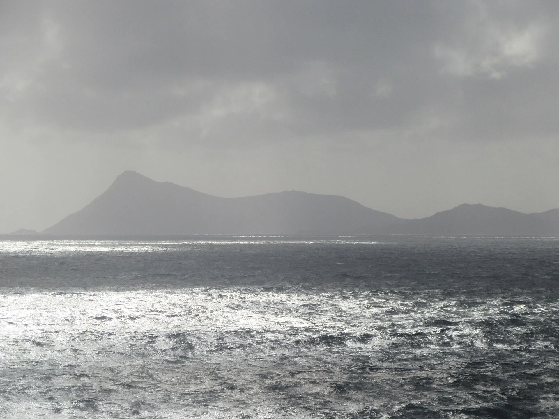 Mar com ondulação e silhueta distante da costa do Cabo de Hornos sob chuva e visibilidade reduzida, sem embarcações em primeiro plano.