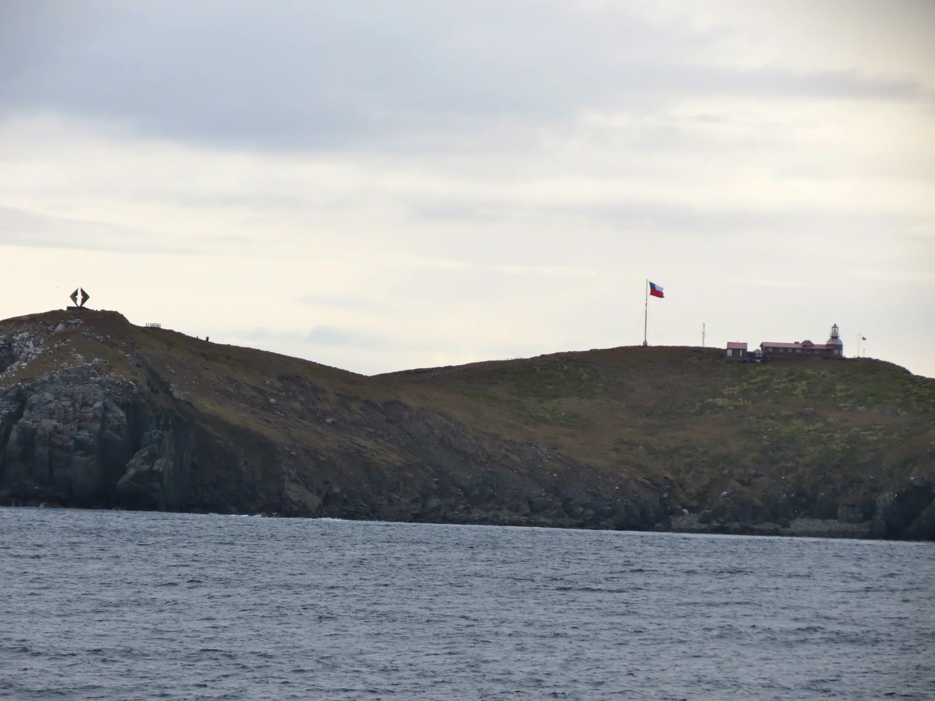 Vista distante do Monumento ao Albatroz, farol e construções da ilha Hornos acima de falésias, com o mar em primeiro plano.