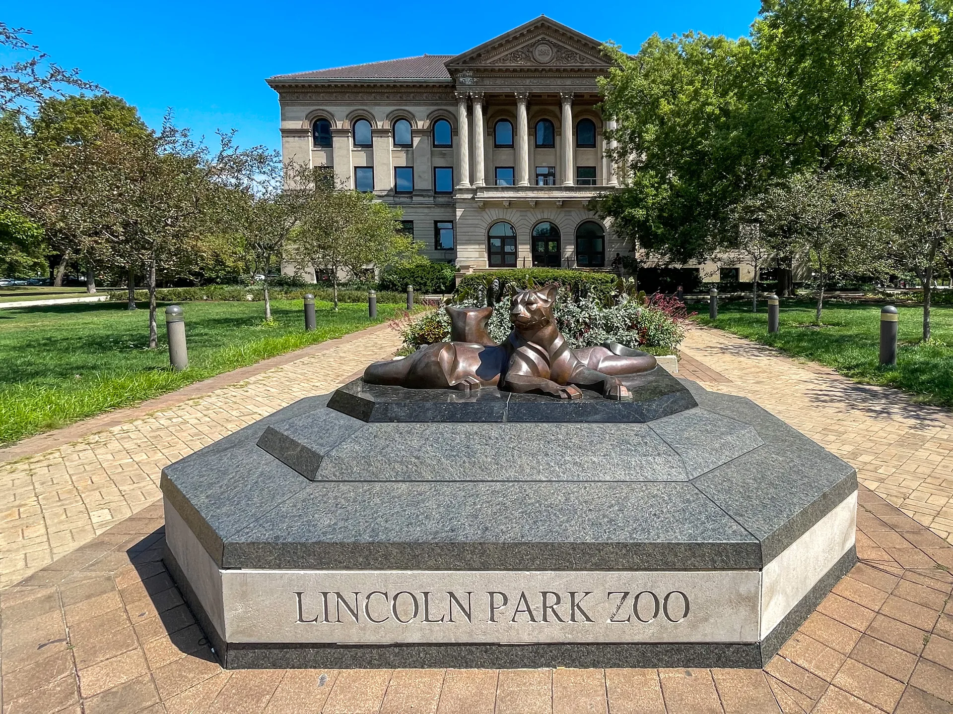 Entrada do Lincoln Park Zoo com escultura de felinos, jardim e edifício histórico sob céu azul.