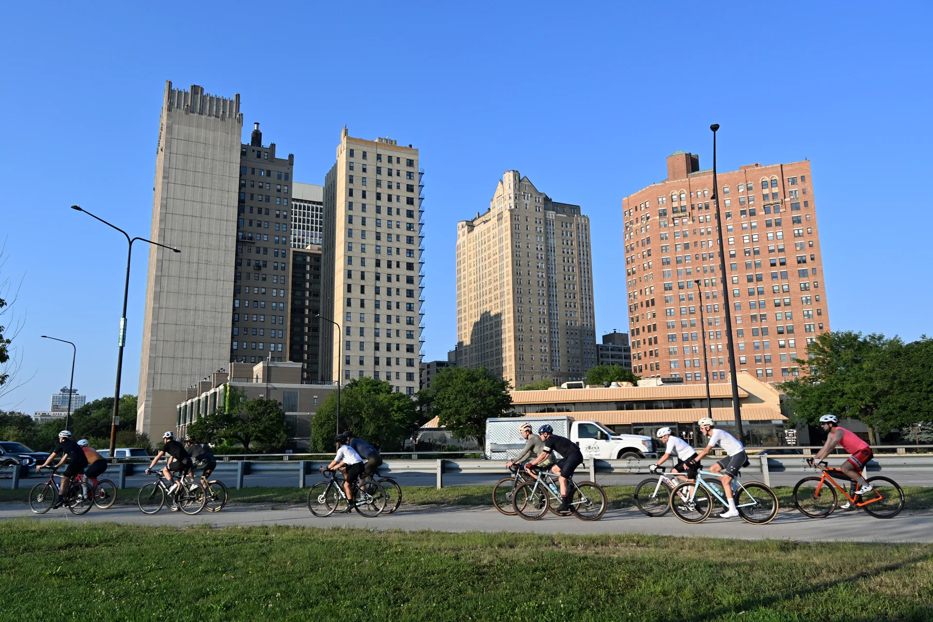 Grupo de ciclistas percorre a Lakefront Trail em Chicago, com gramado, Lake Shore Drive e edifícios de Kenwood ao fundo, em manhã clara.