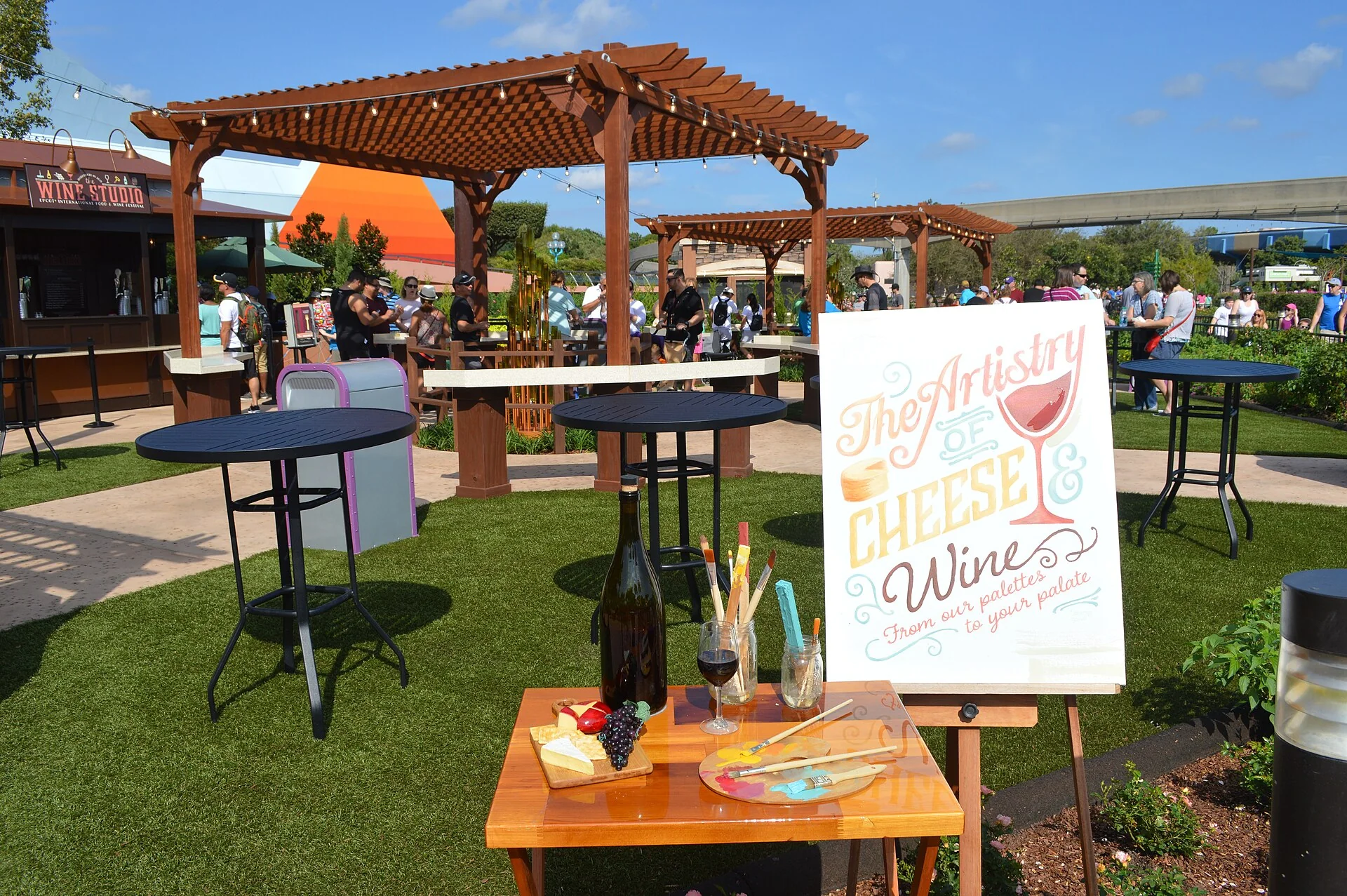 Área externa do EPCOT International Food & Wine Festival em dia claro, com mesas altas, sinalização de queijos e vinhos e visitantes ao fundo.