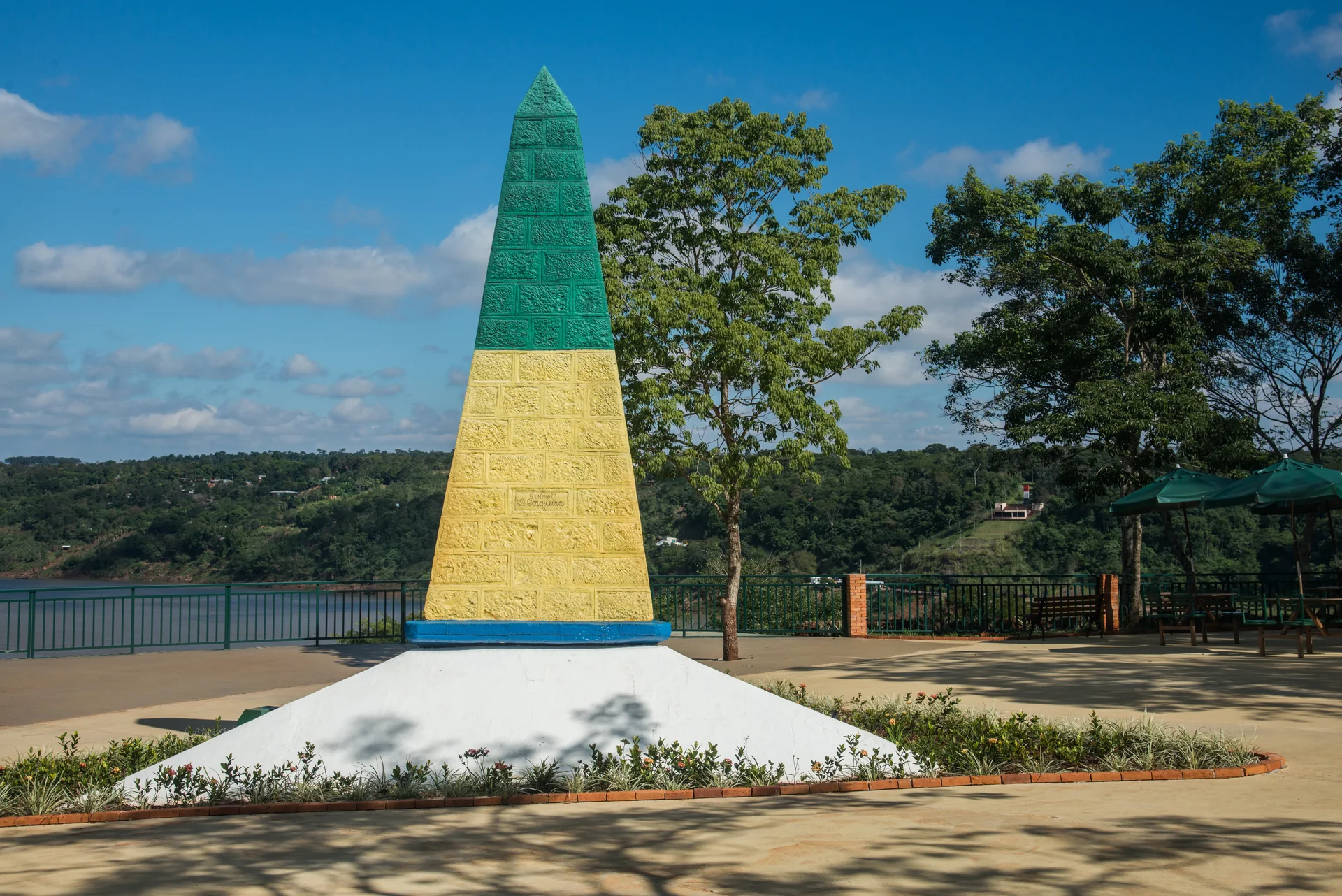 Obelisco verde e amarelo do Marco das Três Fronteiras com os rios e a mata ao fundo.