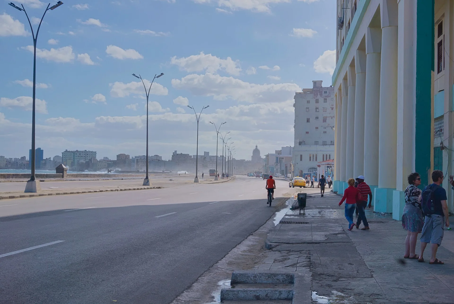 Avenida do Malecón de Havana vista em luz natural, com mar à esquerda, edifícios à direita, pedestres e um ciclista.