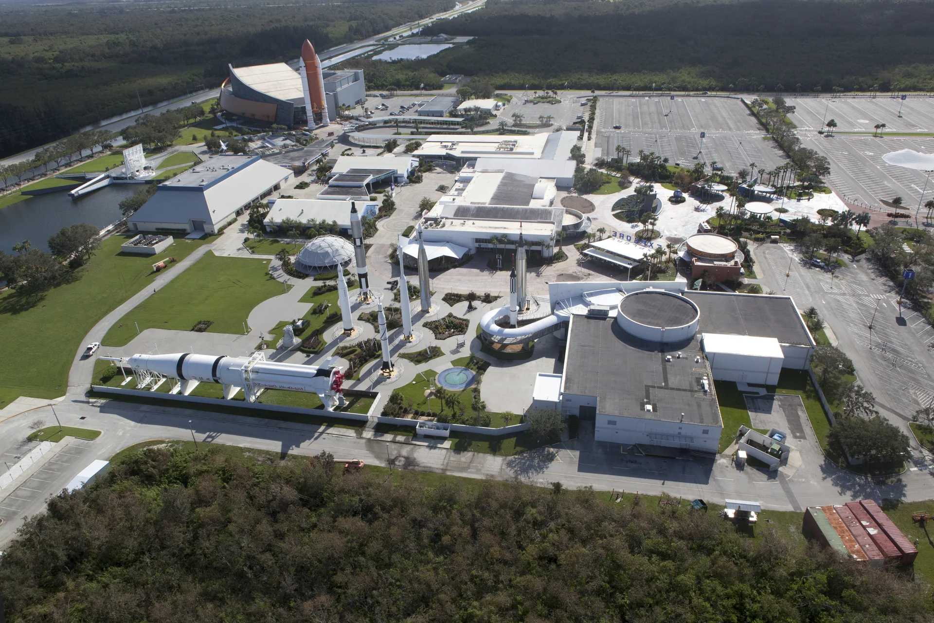 Vista aérea do Kennedy Space Center Visitor Complex, com edifícios, áreas de circulação e o entorno arborizado de Merritt Island.