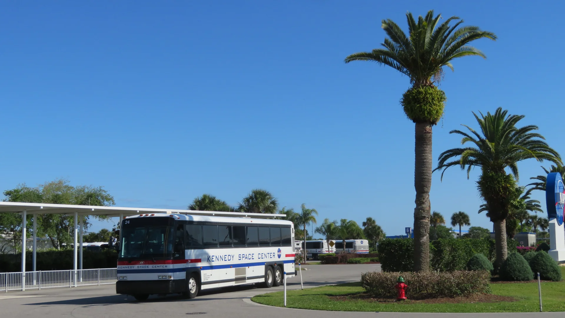 Ônibus de visitantes do Kennedy Space Center parado em área externa arborizada, sob céu azul.