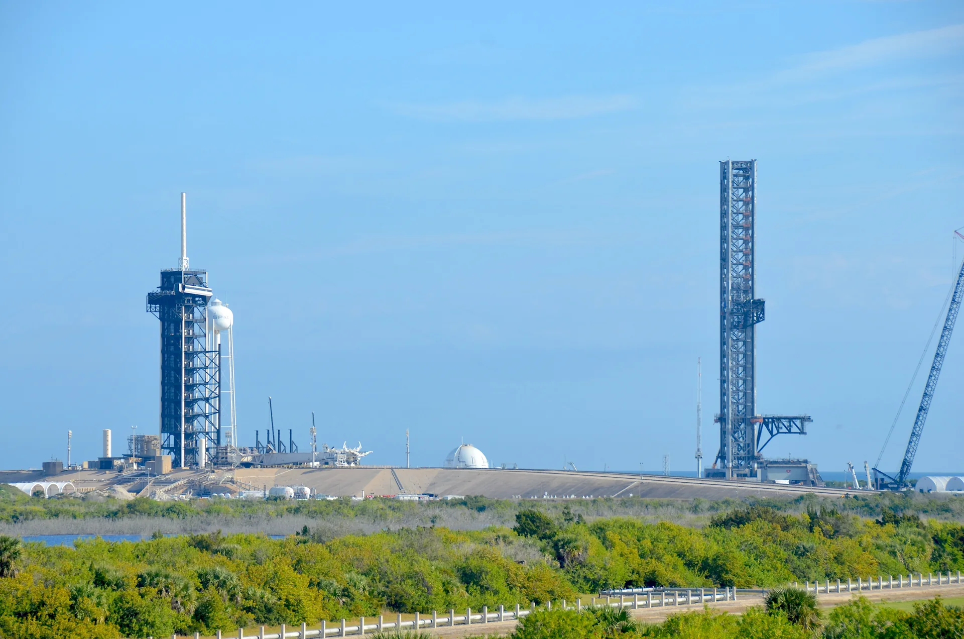 Torres e estruturas do Launch Complex 39A vistas a distância a partir do The Gantry, com vegetação e céu claro em Merritt Island.