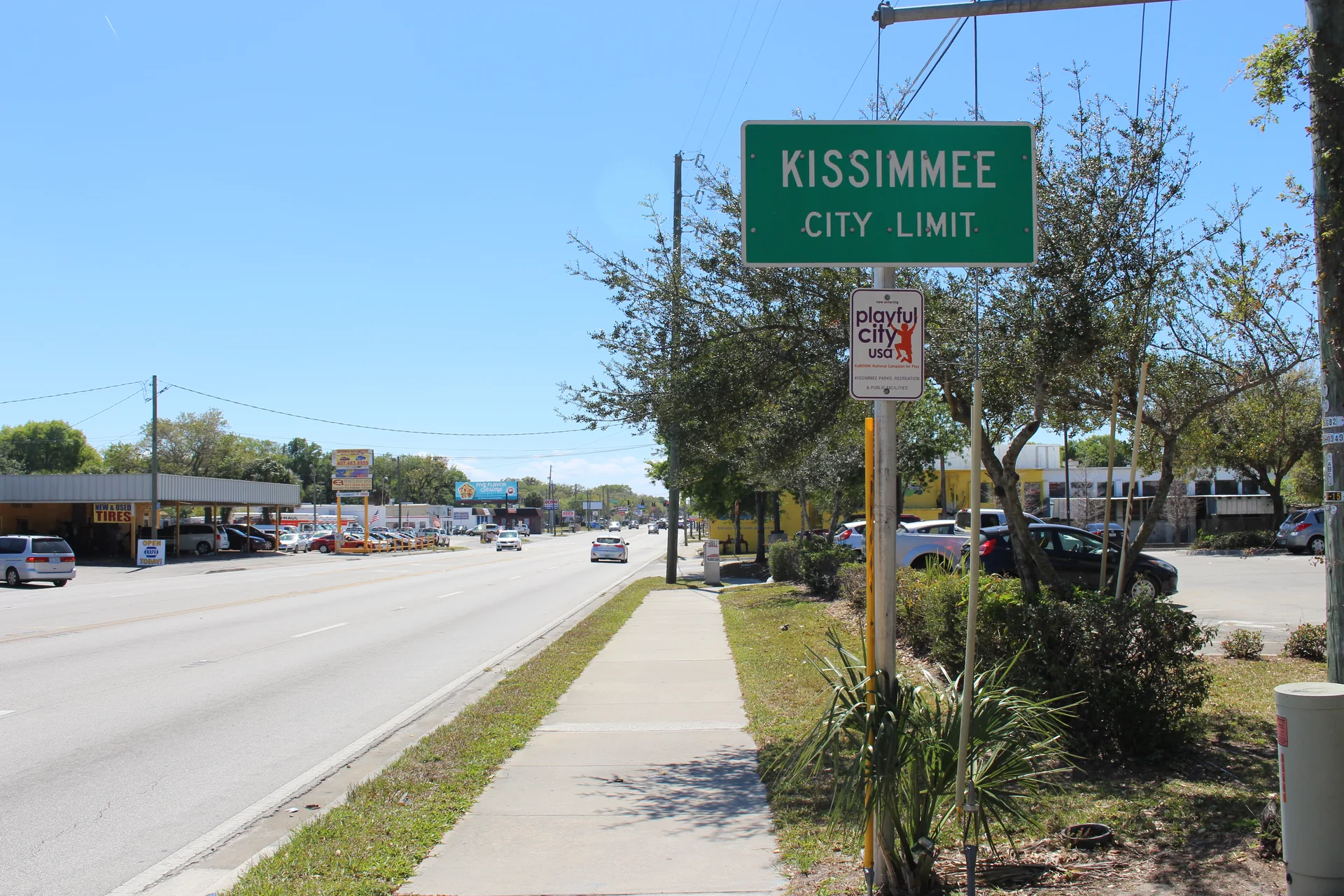 Placa de limite urbano de Kissimmee ao lado de via movimentada, calçada e céu azul.
