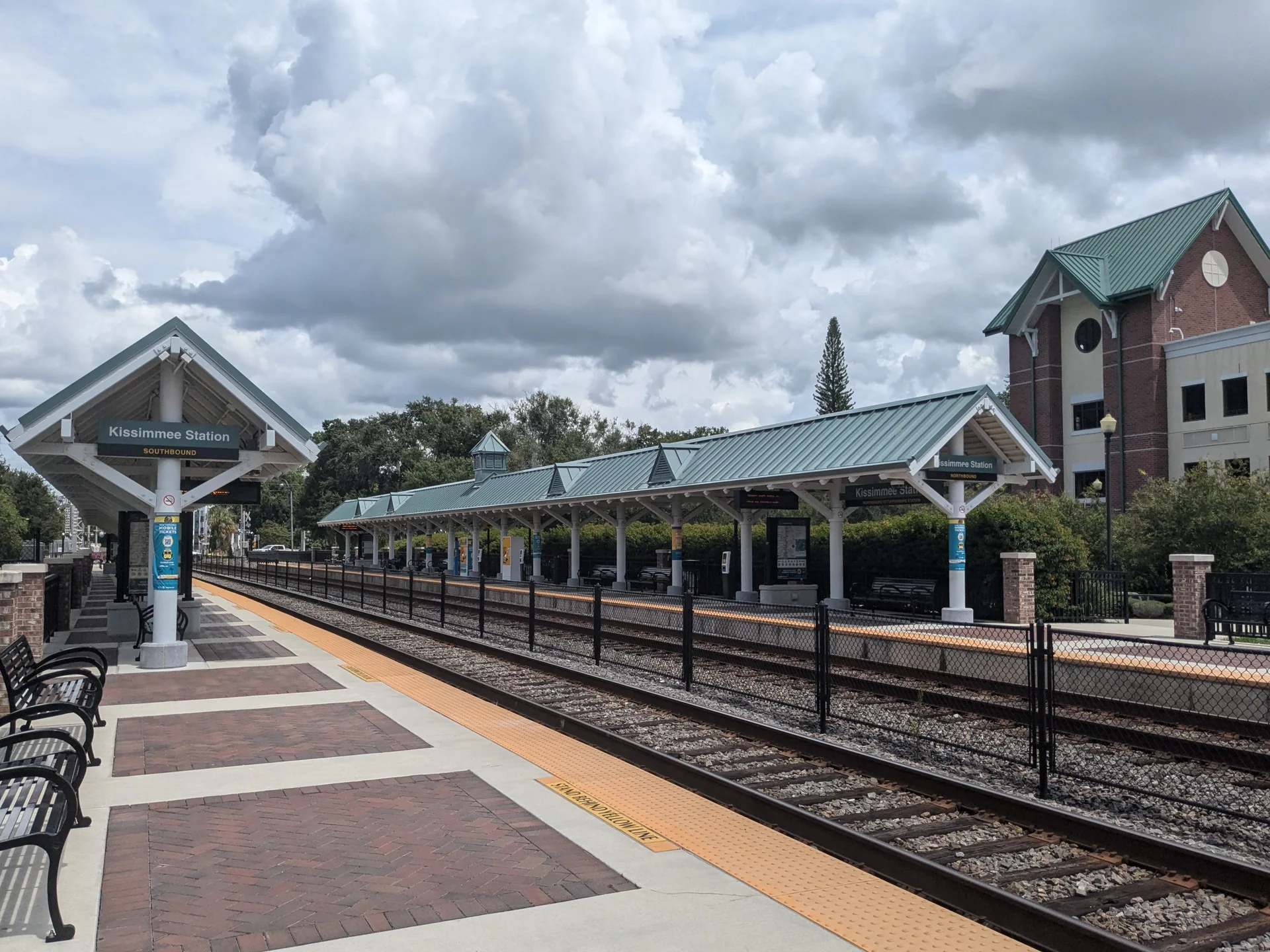 Plataformas do SunRail junto à estação Kissimmee/Amtrak, com cobertura, trilhos e céu nublado.