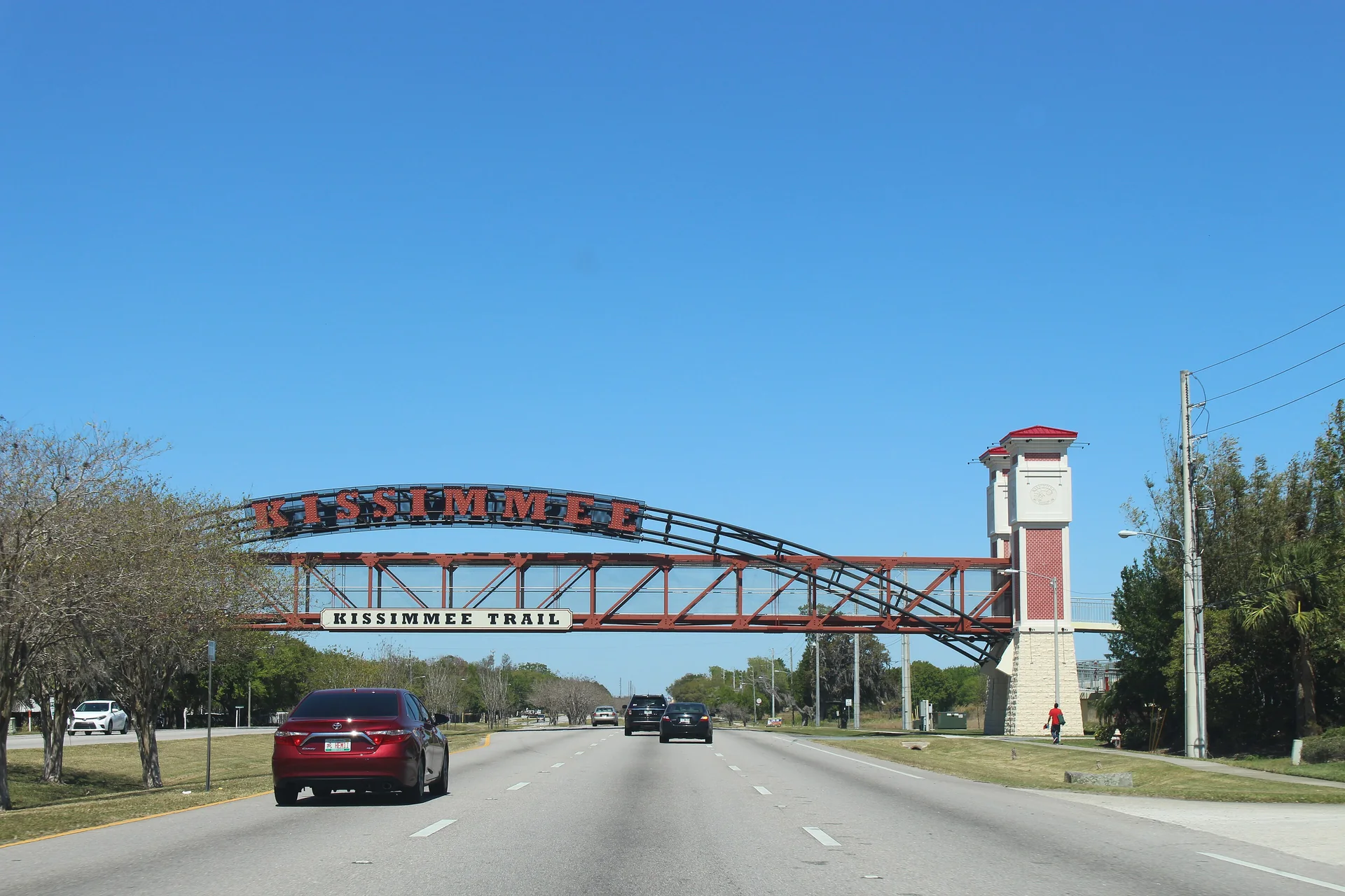 Passarela da Kissimmee Trail sobre a North John Young Parkway, com pista, torre e céu azul.