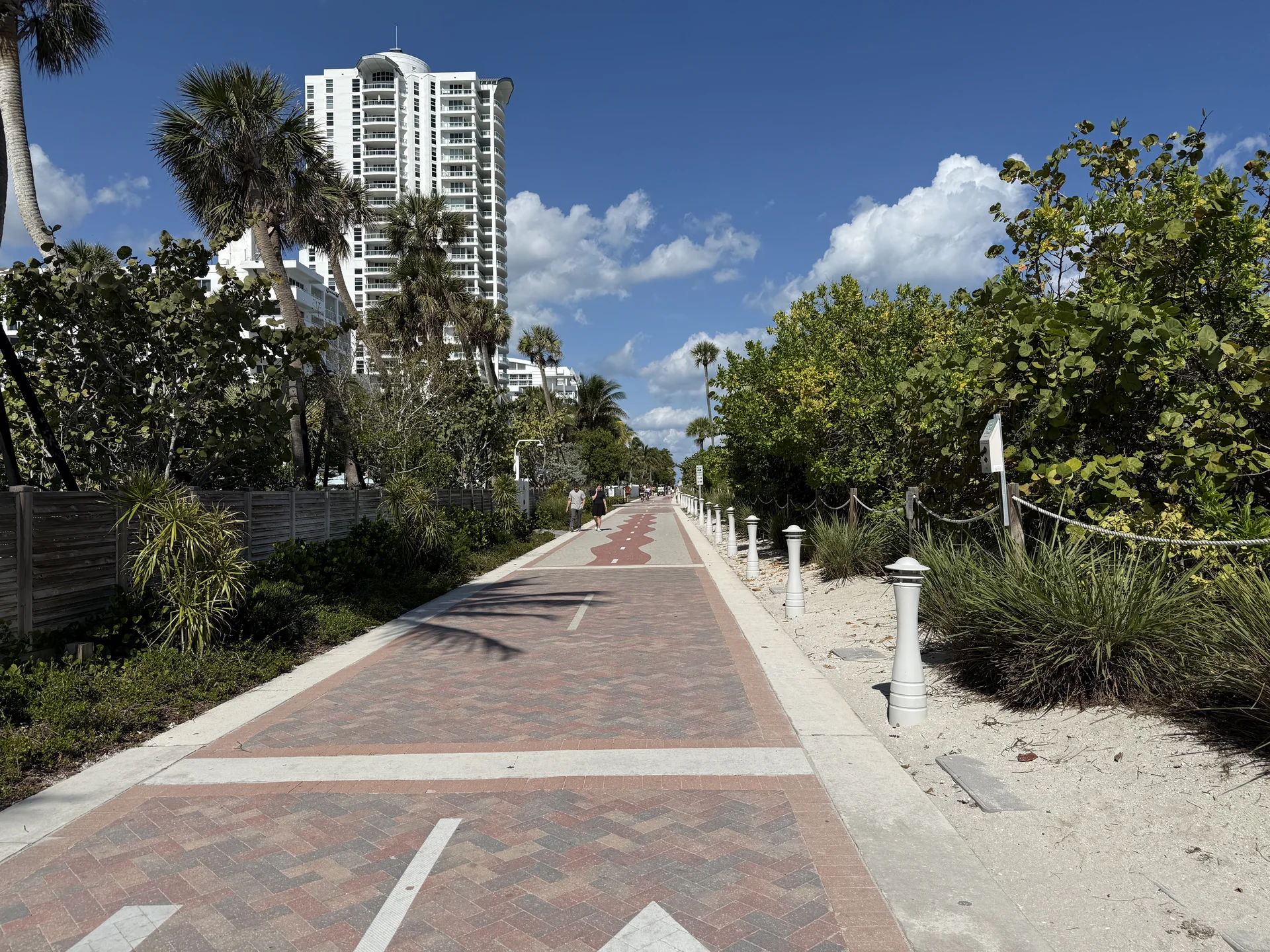 Trecho amplo e sinalizado do Miami Beach Boardwalk perto da 40th Street, com pedestres ao longe, vegetação costeira, palmeiras, prédios e céu azul.
