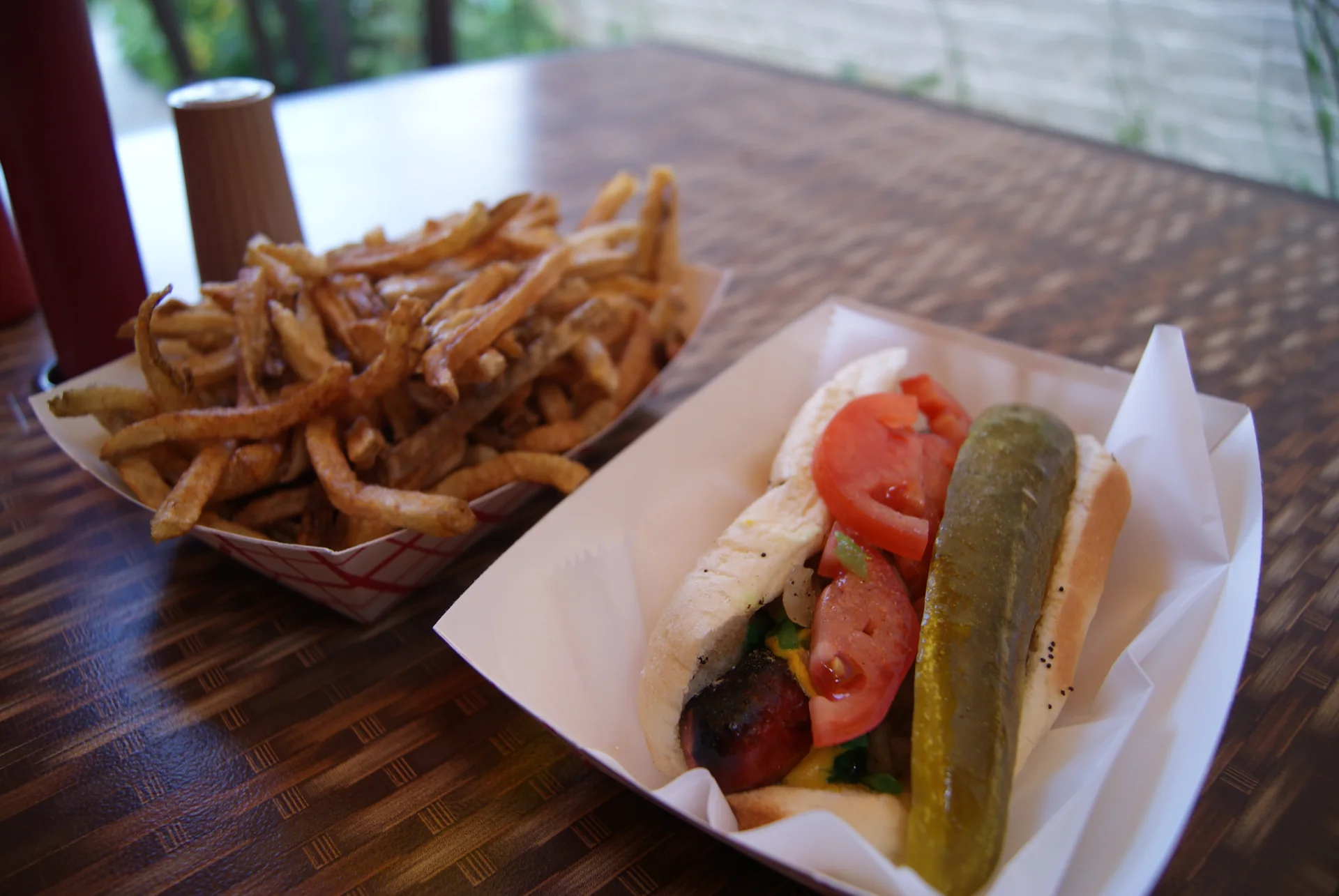 Chicago-style hot dog em bandeja de papel, com picles, tomate e batatas fritas ao lado.