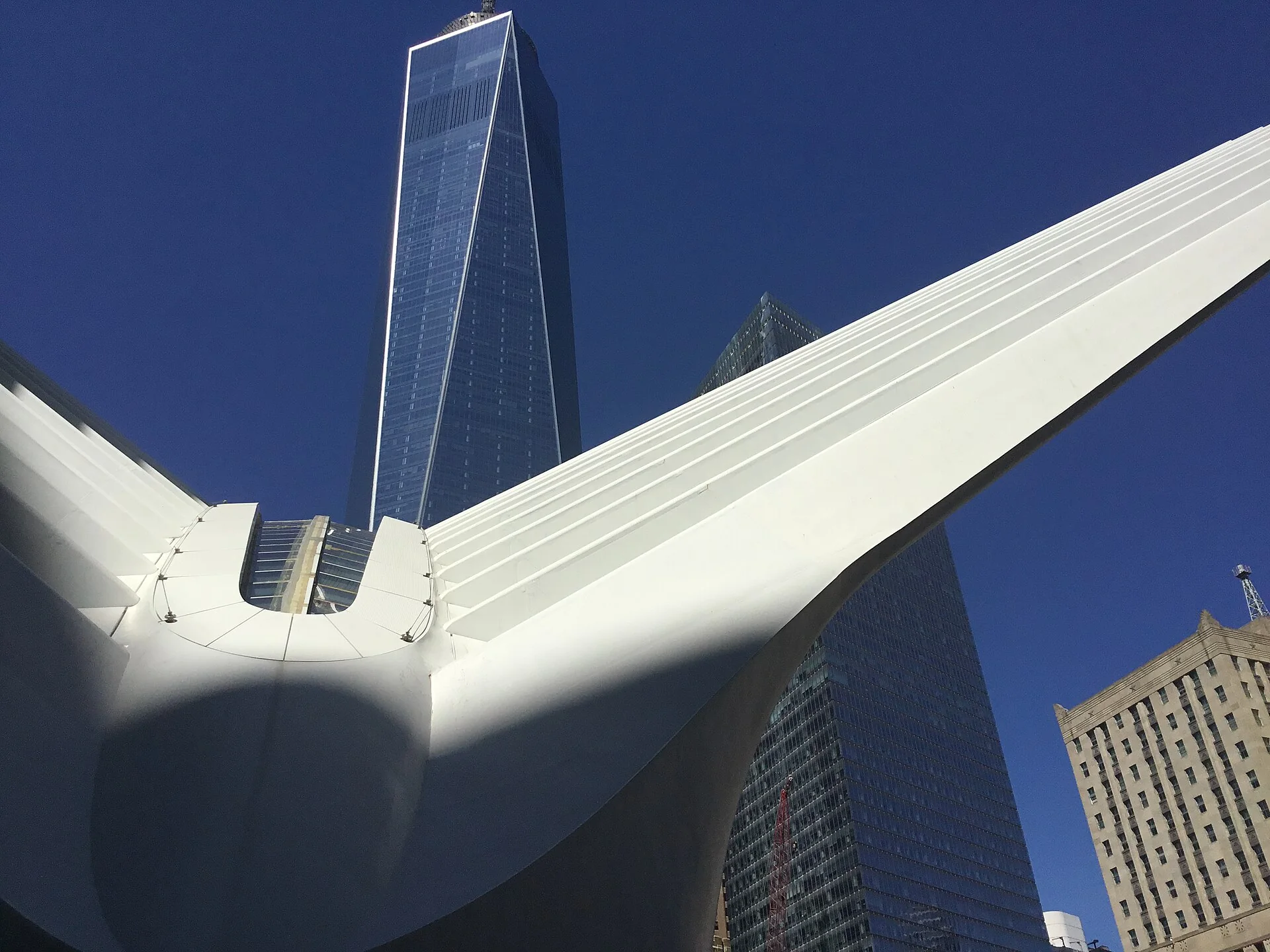One World Trade Center visto ao lado da estrutura branca externa do Oculus, sob céu azul em Lower Manhattan.