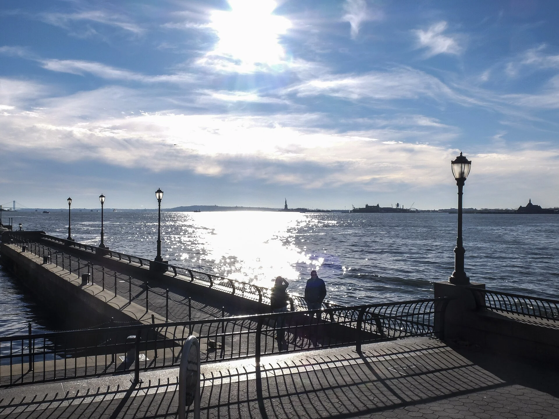 Calçadão de Battery Park City em luz diurna, com postes, pessoas, água e a Estátua da Liberdade ao fundo.