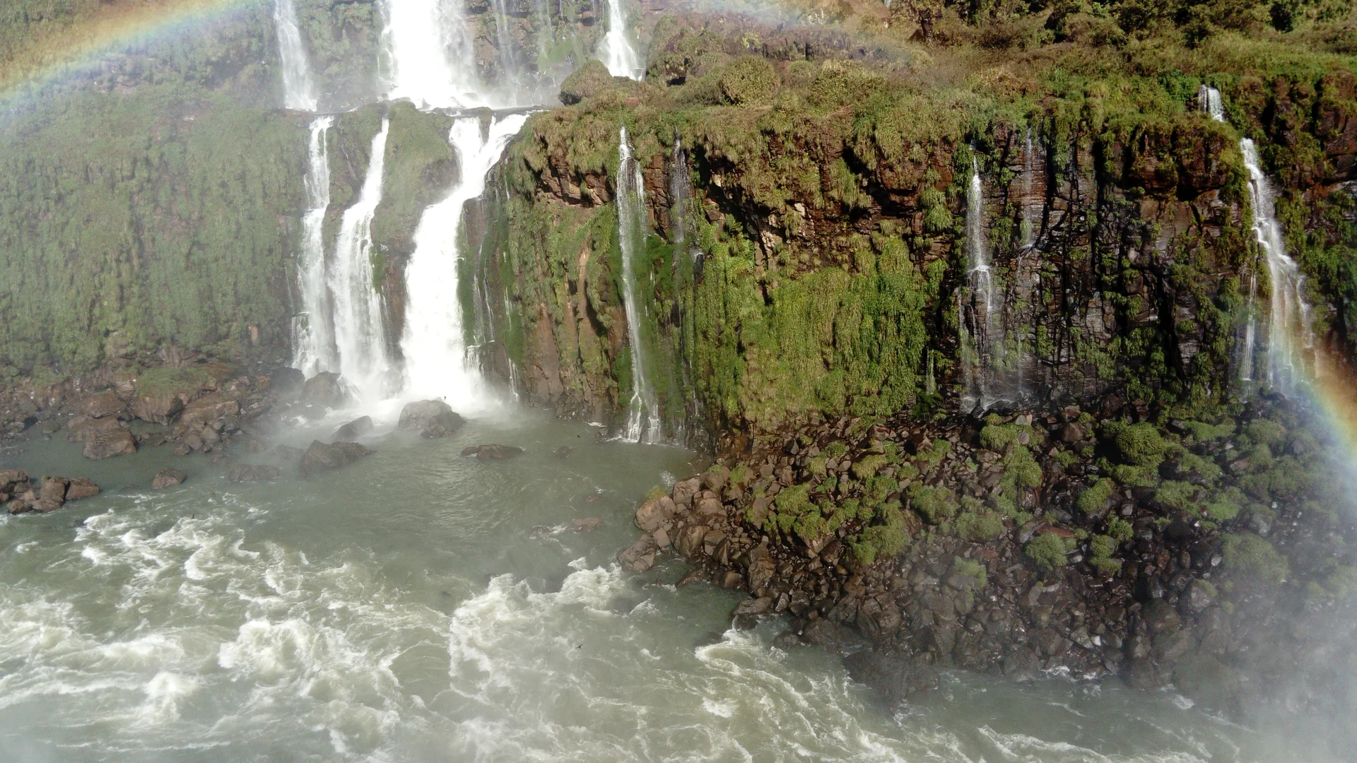 Detalhe das quedas das Cataratas do Iguaçu com arco-íris e rochas cobertas por vegetação.