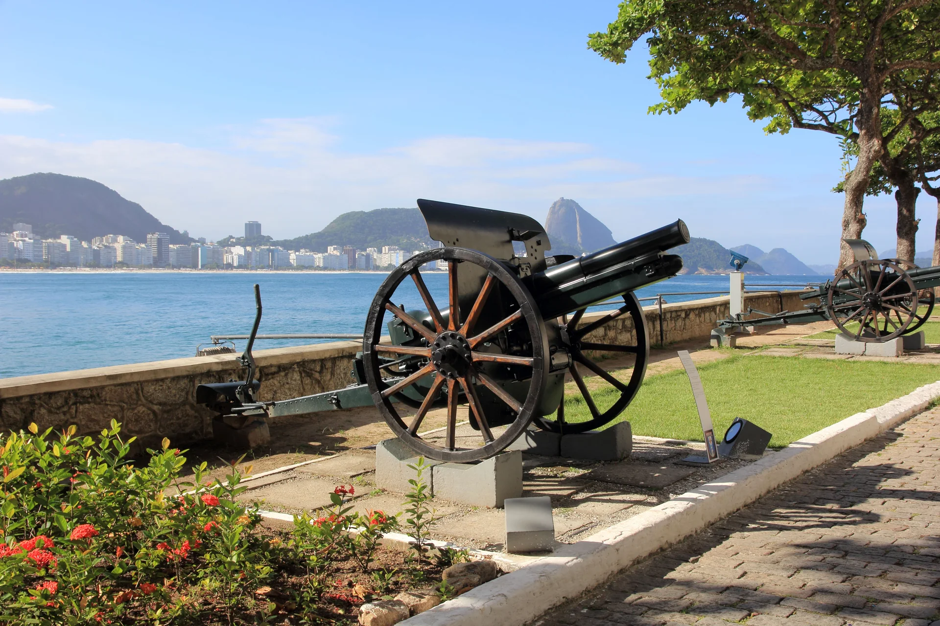 Canhões do Forte de Copacabana sob céu azul, com a Praia de Copacabana, o mar e o Pão de Açúcar ao fundo; imagem contextual, não representa o parceiro.