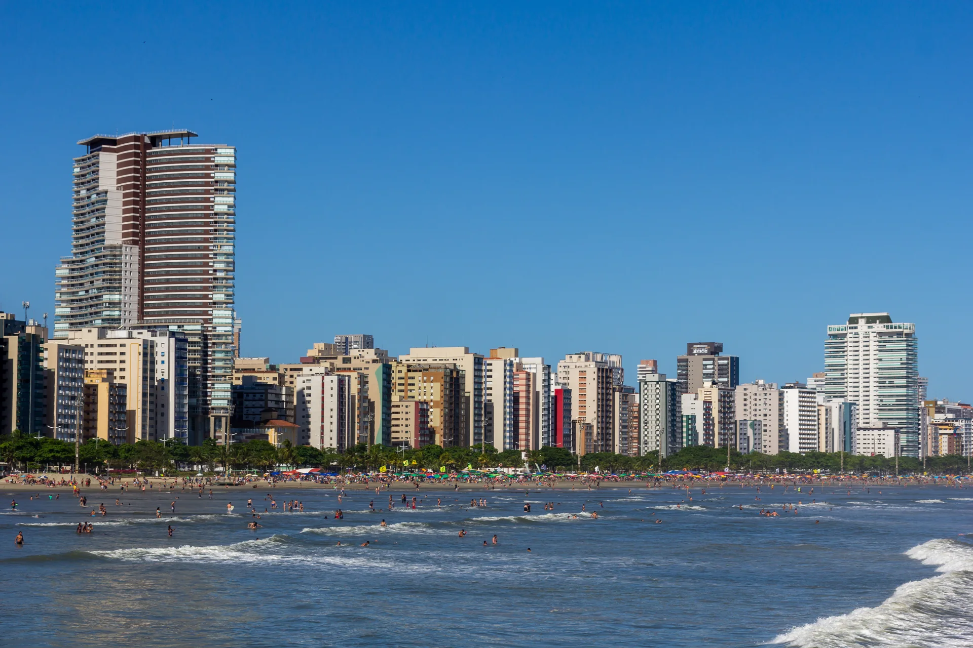 Orla ensolarada de Santos, com praia, mar, banhistas e edifícios sob céu azul.