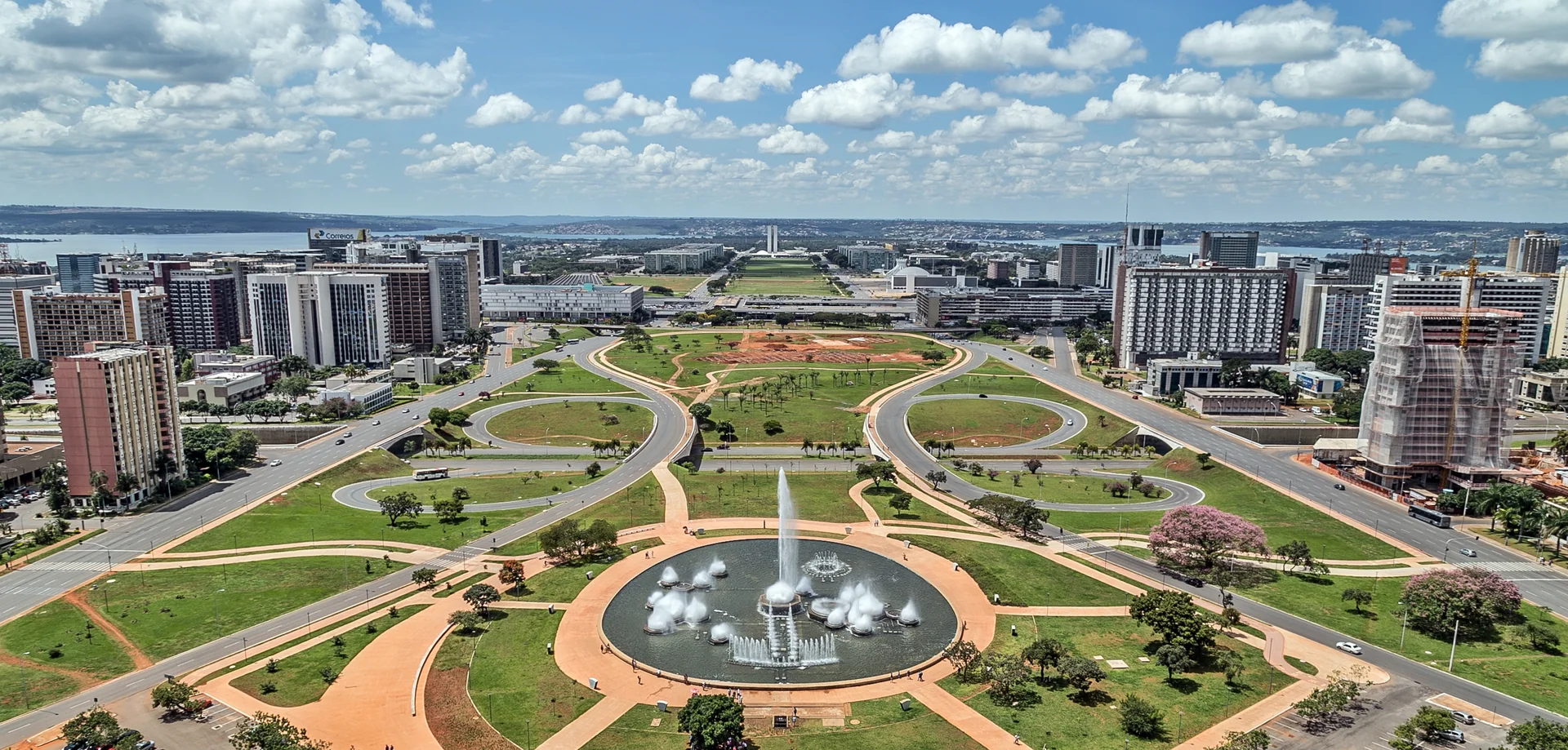 Vista diurna do Eixo Monumental de Brasília, com fontes, vias, gramados e céu claro.