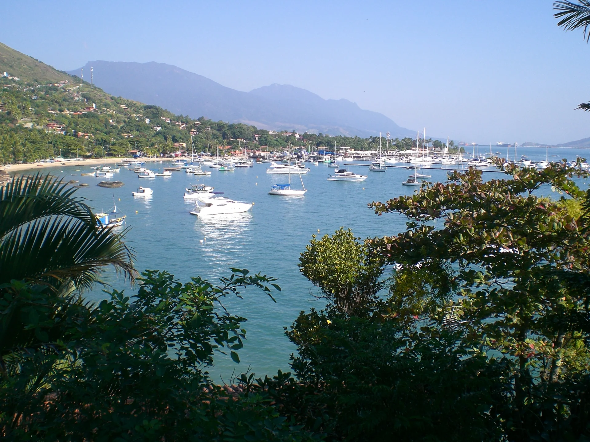 Imagem de contexto de baía ensolarada em Ilhabela com embarcações e vegetação; não representa o parceiro, a propriedade ou os serviços do Hostel da Vila Ilhabela.