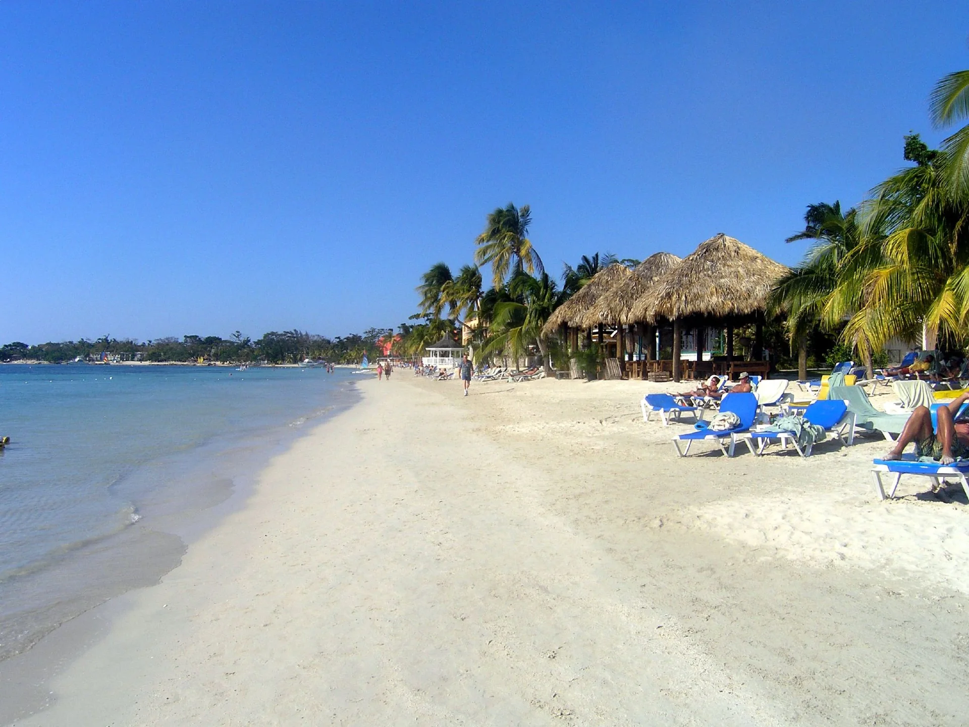Praia ensolarada no Sandals Negril, Jamaica, com palmeiras, espreguiçadeiras e mar ao fundo.