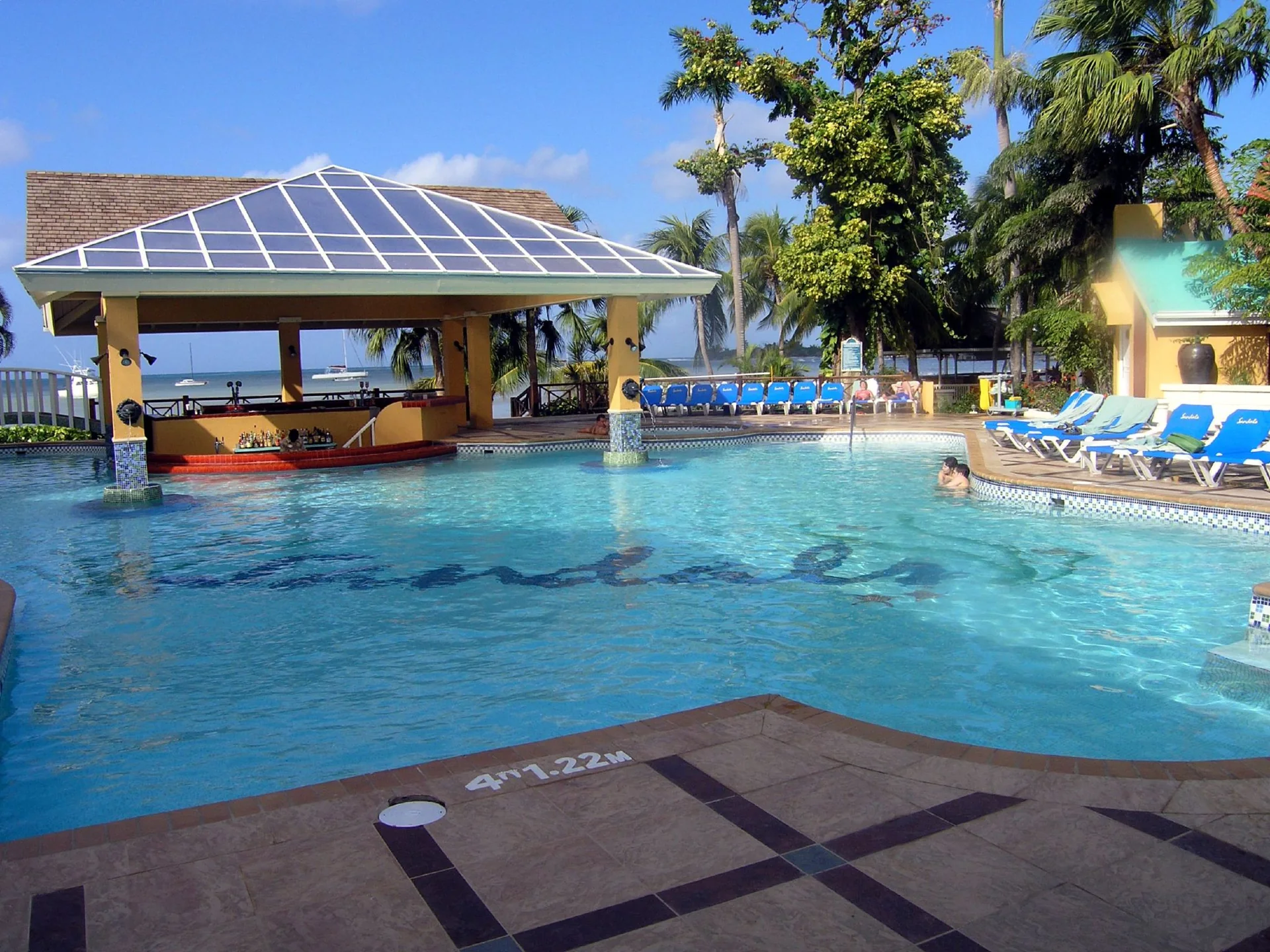 Piscina externa ensolarada no Sandals Negril, Jamaica, com bar integrado e palmeiras.