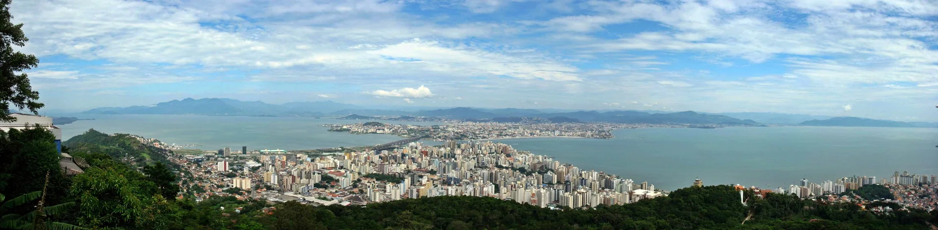 Contexto do destino: panorama colorido e ensolarado de Florianópolis, visto do continente sob céu azul; não representa a Seazone nem acomodações.