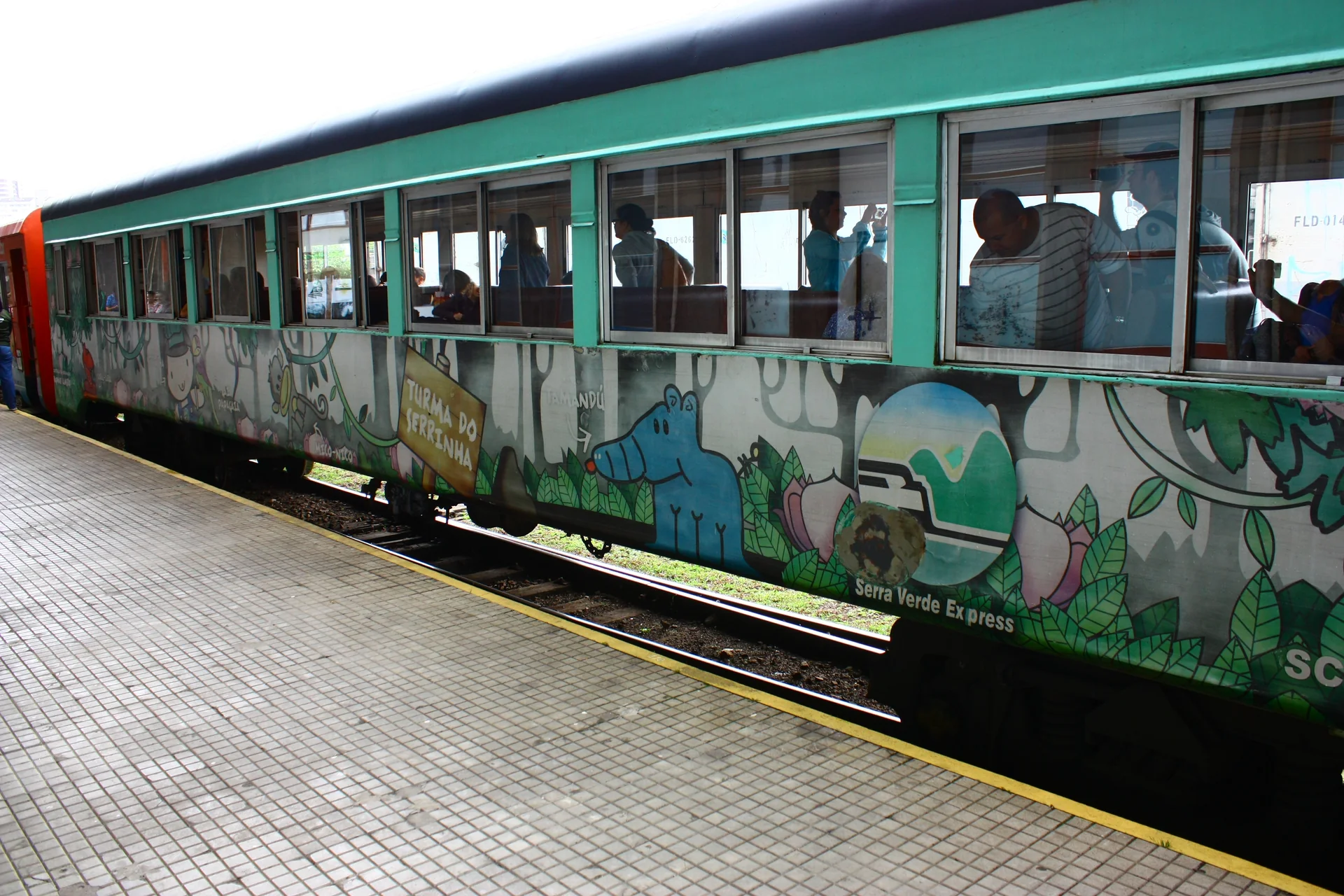Vagão do trem turístico parado junto à plataforma, com passageiros visíveis pelas janelas.