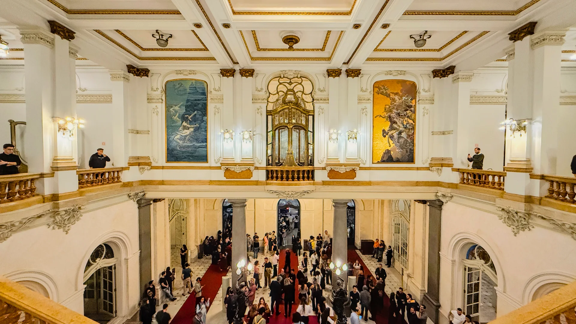 Vista interna do saguão ornamentado do Theatro Municipal de São Paulo, com escadaria e visitantes.